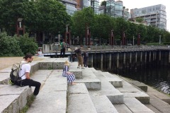 7 people are widely spaced, standing and sitting on concrete steps that lead down to the water at False Creek in Vancouver.